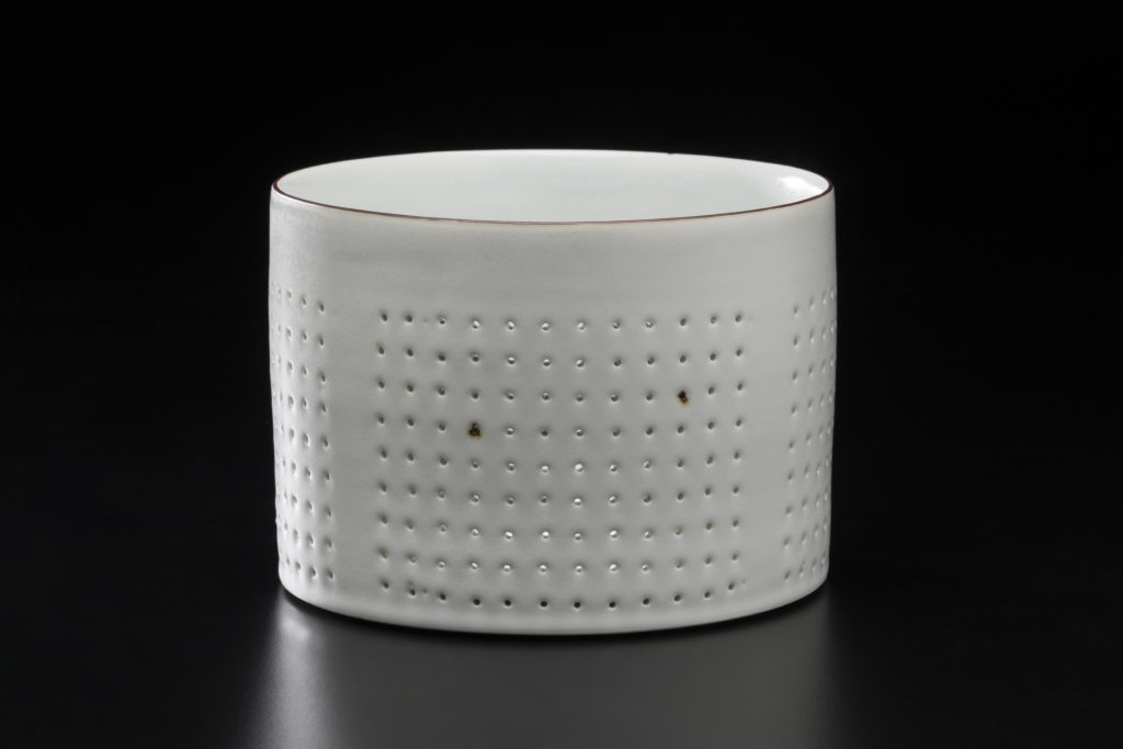 A white cylindrical box in white porcelain decorated with rows of small holes. The top edge and two holes show delicate rust-like brown stains.